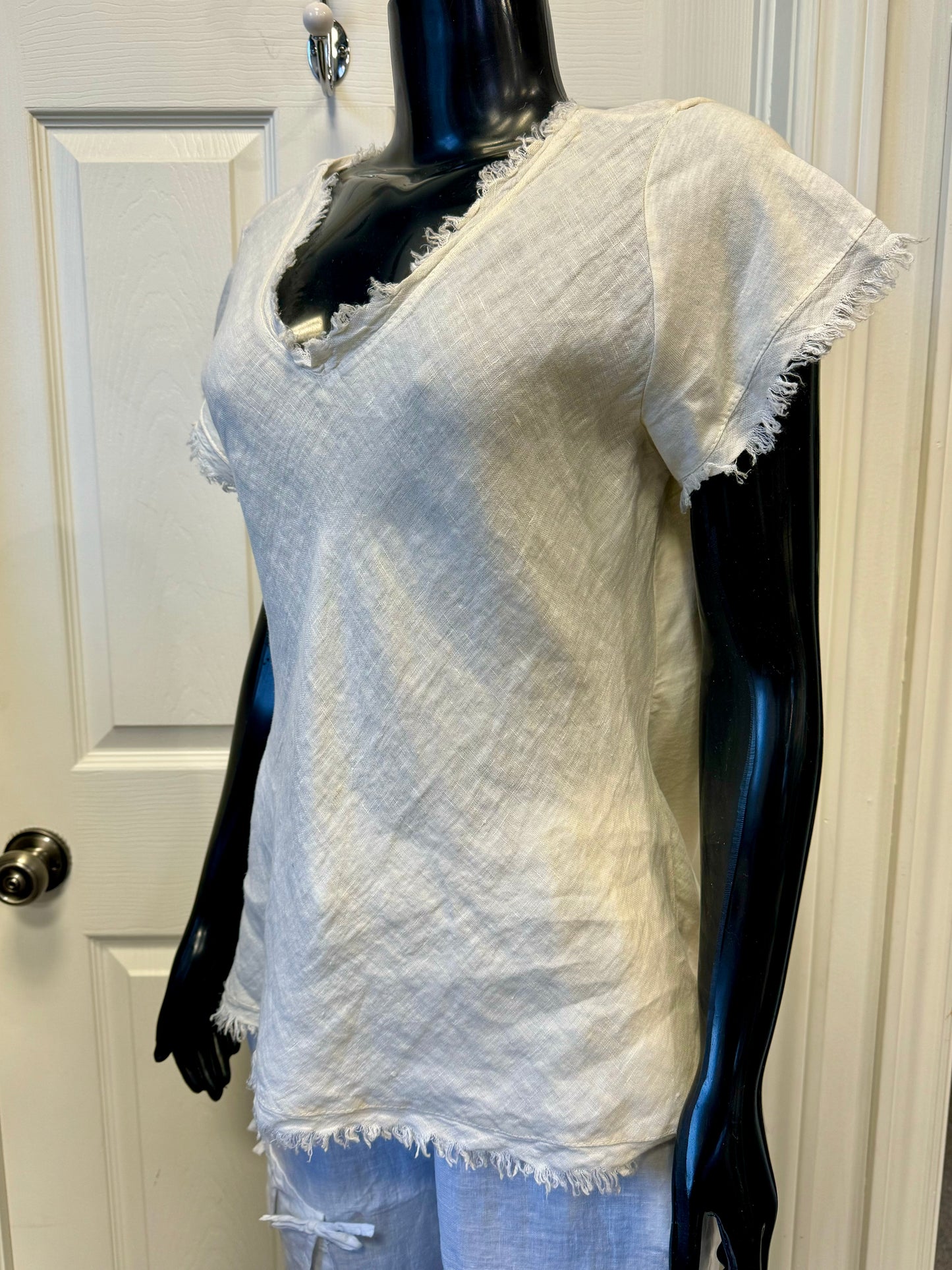 A mannequin showcases the LINEN TOP VANILLA- ETERNELLE by ETERNELLE, featuring a light beige, short-sleeved, V-neck design with frayed edges. The 100% linen fabric has a textured, woven appearance. The vanilla-hued top is paired with matching pants and displayed against a white-paneled door background.