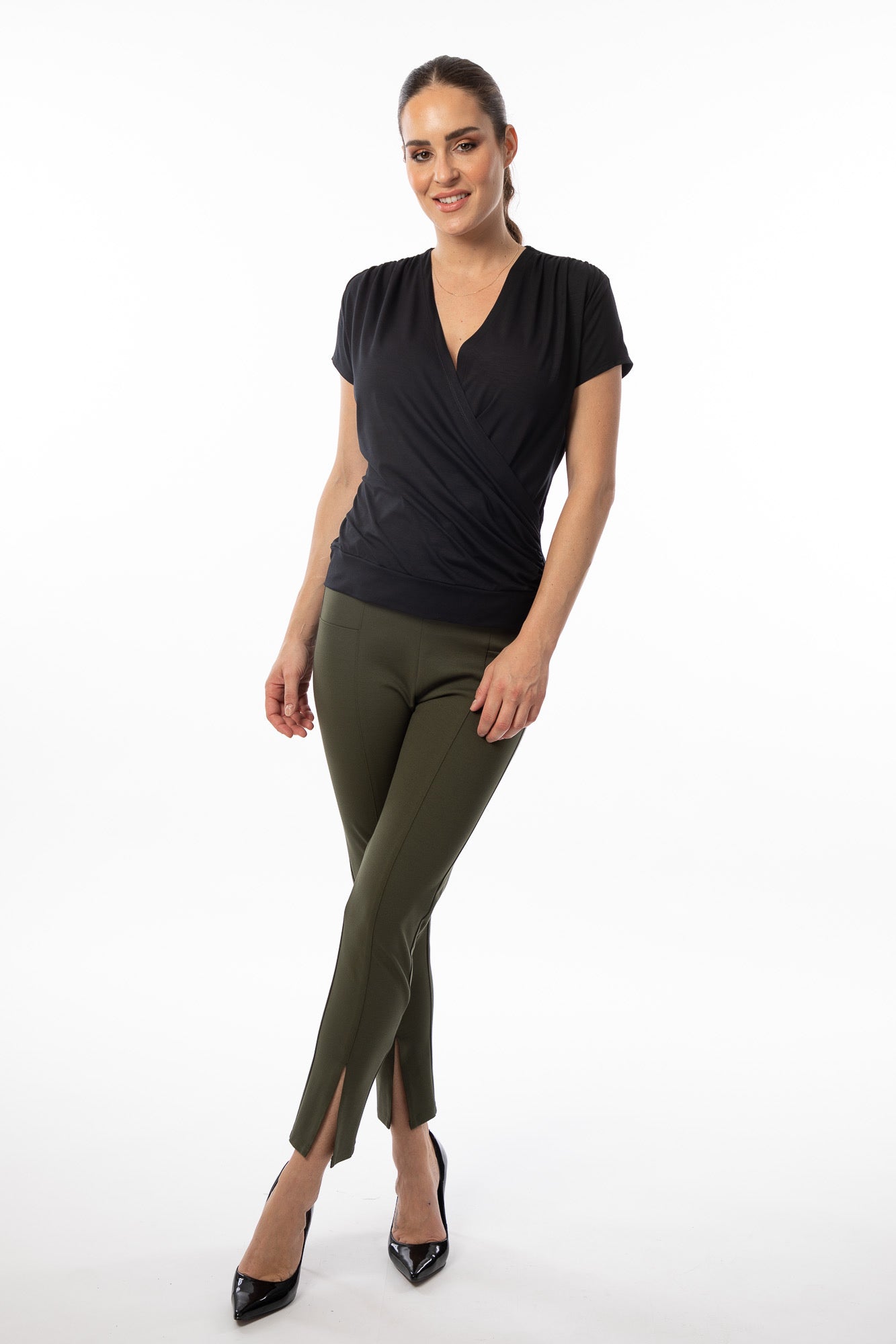 A woman posed against a white background, wearing an elegant black V-neck blouse (Bali Top 8367 - BLACK by Bali Corp) and olive green pants with a front slit at the ankles. She is also wearing black high heels and has her brown hair tied back. Smiling, she stands with one leg slightly crossed in front for a stylish fit.