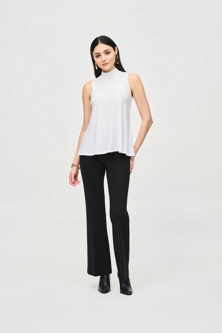 A woman with long dark hair stands against a plain background. She is wearing a Joseph Ribkoff Silky Knit Fit and Flare Sleeveless Top 243253, black flared pants, and black ankle boots. She has minimal jewelry and is striking a relaxed, confident pose with a slight smile.