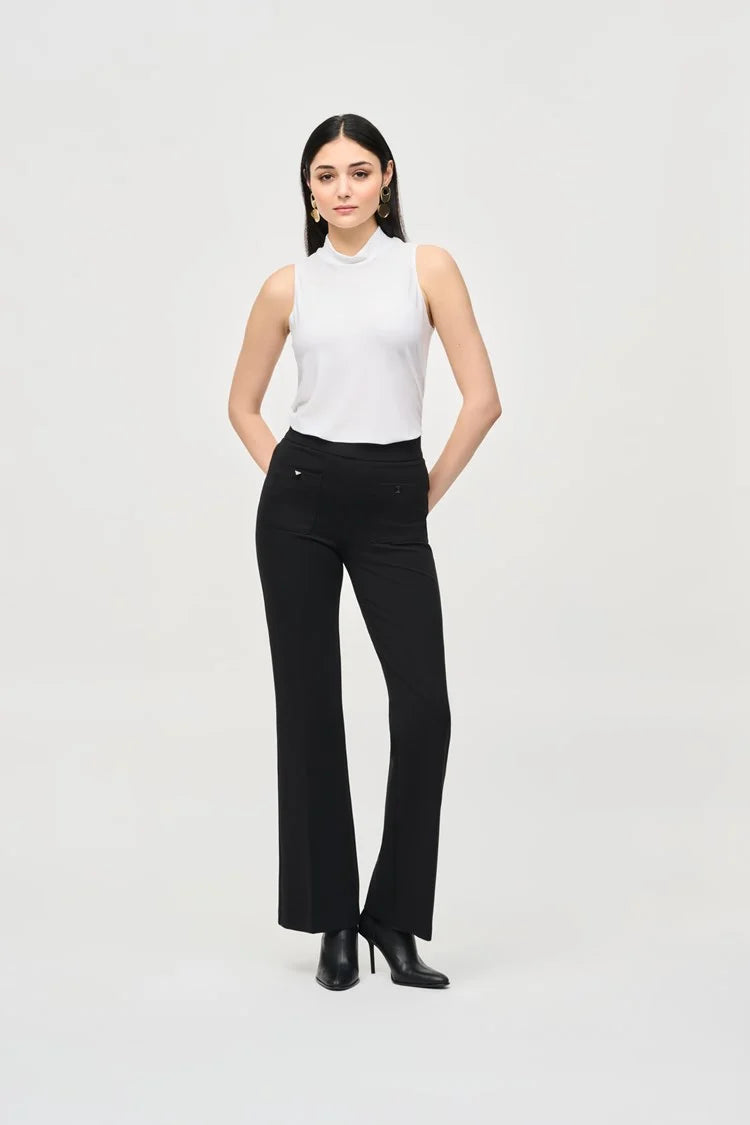 A woman with long dark hair stands against a plain background. She is wearing a Joseph Ribkoff Silky Knit Fit and Flare Sleeveless Top 243253, black flared pants, and black ankle boots. She has minimal jewelry and is striking a relaxed, confident pose with a slight smile.