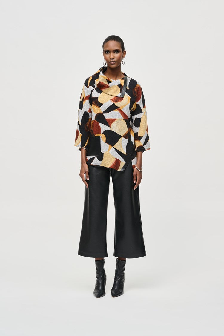 A person stands against a plain background, wearing the Joseph Ribkoff Sweater Knit Abstract Print Boxy Top (model number 243291) featuring geometric patterns in shades of black, yellow, white, and rust. The top has dolman sleeves and is paired with black wide-legged pants and black ankle boots. The person has short hair and complements the outfit with large hoop earrings.