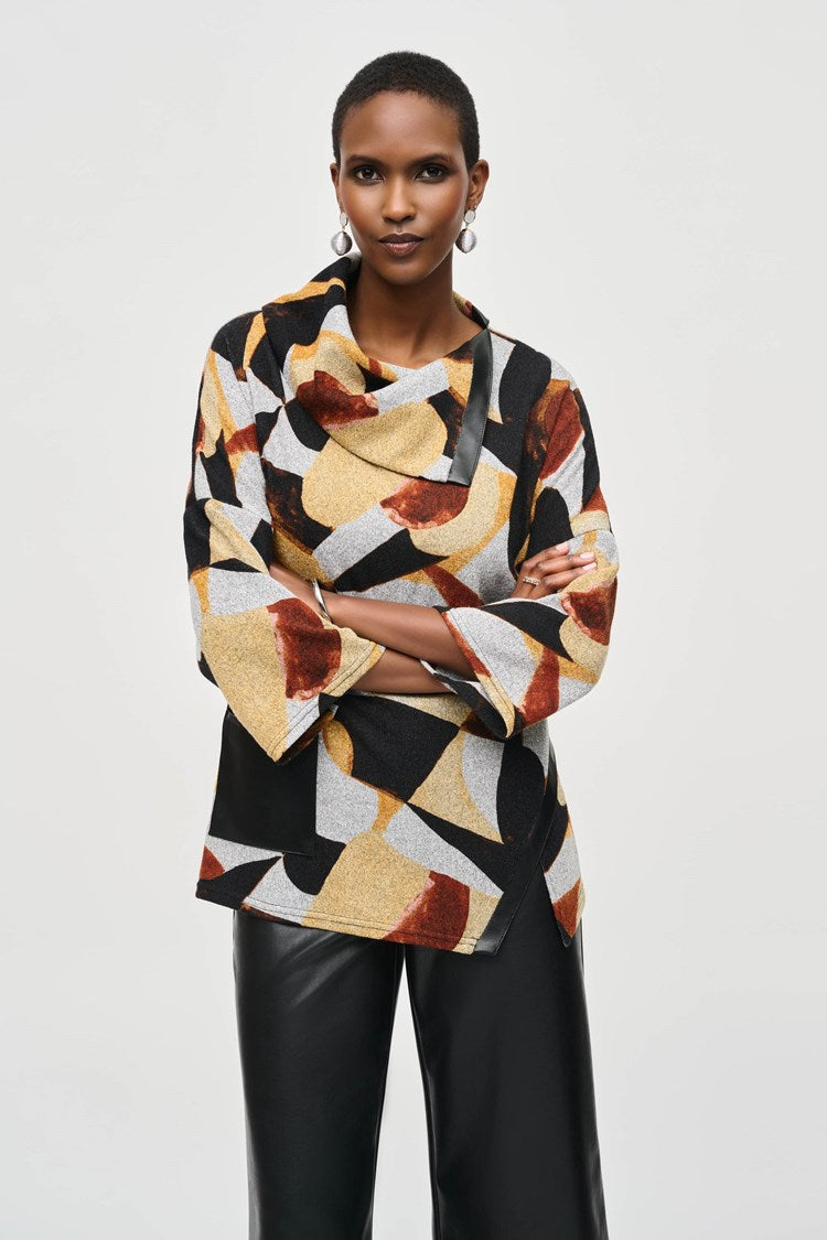 A person stands against a plain background, wearing the Joseph Ribkoff Sweater Knit Abstract Print Boxy Top (model number 243291) featuring geometric patterns in shades of black, yellow, white, and rust. The top has dolman sleeves and is paired with black wide-legged pants and black ankle boots. The person has short hair and complements the outfit with large hoop earrings.