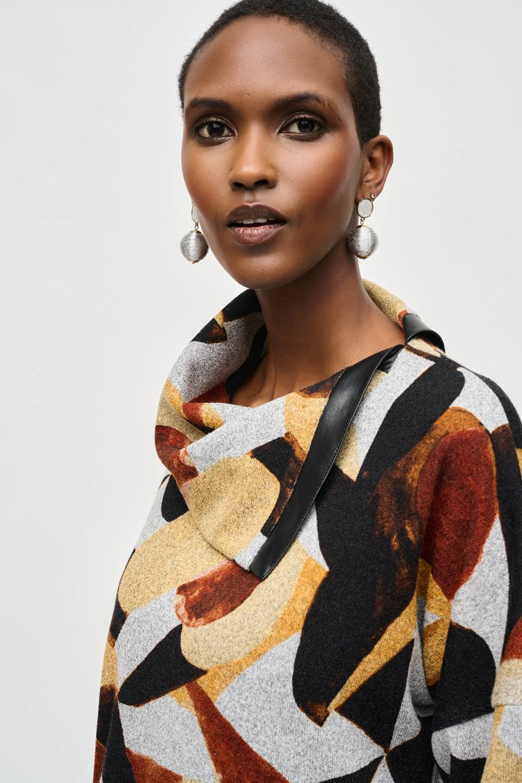 A person stands against a plain background, wearing the Joseph Ribkoff Sweater Knit Abstract Print Boxy Top (model number 243291) featuring geometric patterns in shades of black, yellow, white, and rust. The top has dolman sleeves and is paired with black wide-legged pants and black ankle boots. The person has short hair and complements the outfit with large hoop earrings.