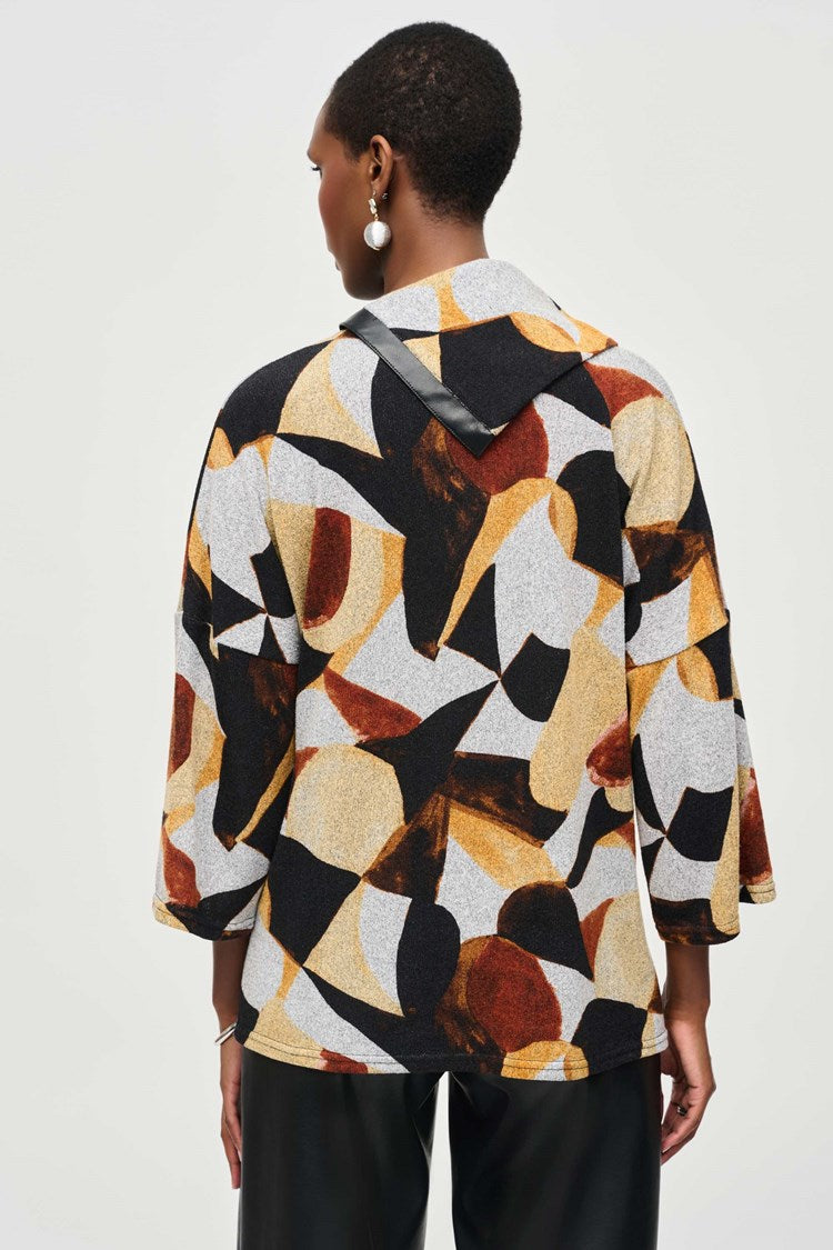 A person stands against a plain background, wearing the Joseph Ribkoff Sweater Knit Abstract Print Boxy Top (model number 243291) featuring geometric patterns in shades of black, yellow, white, and rust. The top has dolman sleeves and is paired with black wide-legged pants and black ankle boots. The person has short hair and complements the outfit with large hoop earrings.