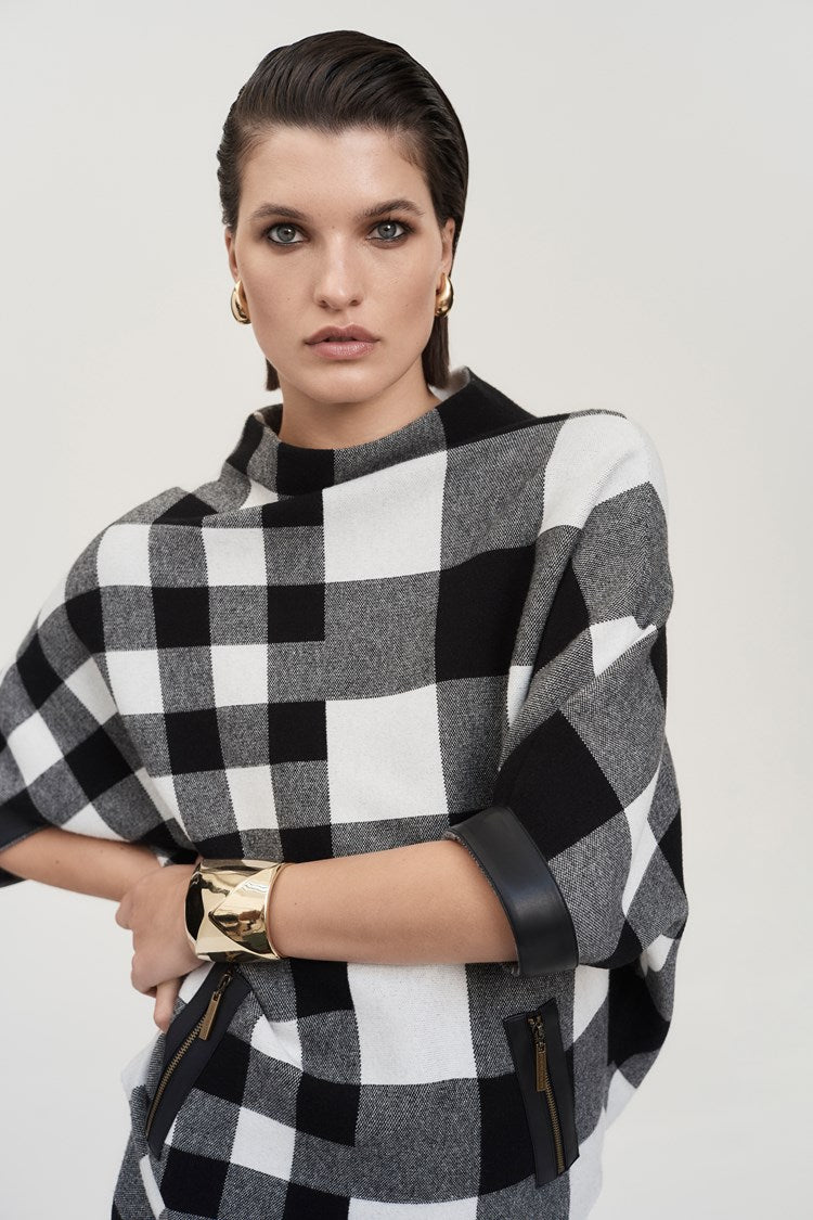 A woman is dressed in the Joseph Ribkoff Plaid Jacquard Knit Sweater 243946 and a matching skirt with leatherette trim. She is standing against a plain white background, wearing black heeled boots and accessorized with earrings and a bracelet. She has long dark hair and is posing confidently.