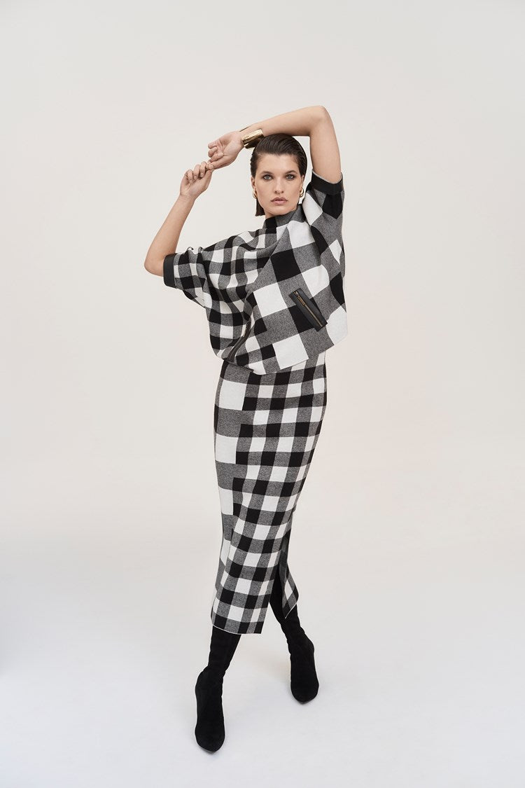 A woman is dressed in the Joseph Ribkoff Plaid Jacquard Knit Sweater 243946 and a matching skirt with leatherette trim. She is standing against a plain white background, wearing black heeled boots and accessorized with earrings and a bracelet. She has long dark hair and is posing confidently.