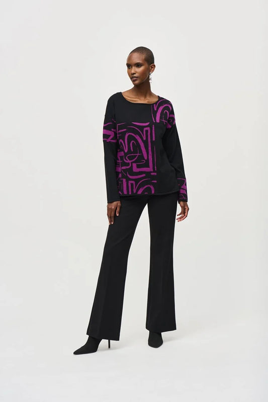 A person stands confidently against a white background wearing a stylish Jacquard Knit Abstract Print Top 243971 by Joseph Ribkoff. They are dressed in a black long-sleeve top featuring vibrant purple geometric patterns on one side and black flared pants. The cozy fabric drapes beautifully as they pose with one hand on their hip.