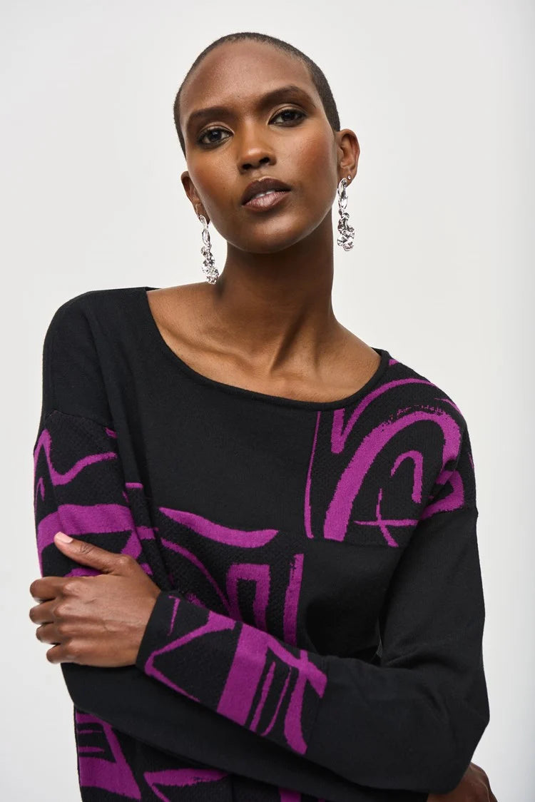 A person stands confidently against a white background wearing a stylish Jacquard Knit Abstract Print Top 243971 by Joseph Ribkoff. They are dressed in a black long-sleeve top featuring vibrant purple geometric patterns on one side and black flared pants. The cozy fabric drapes beautifully as they pose with one hand on their hip.