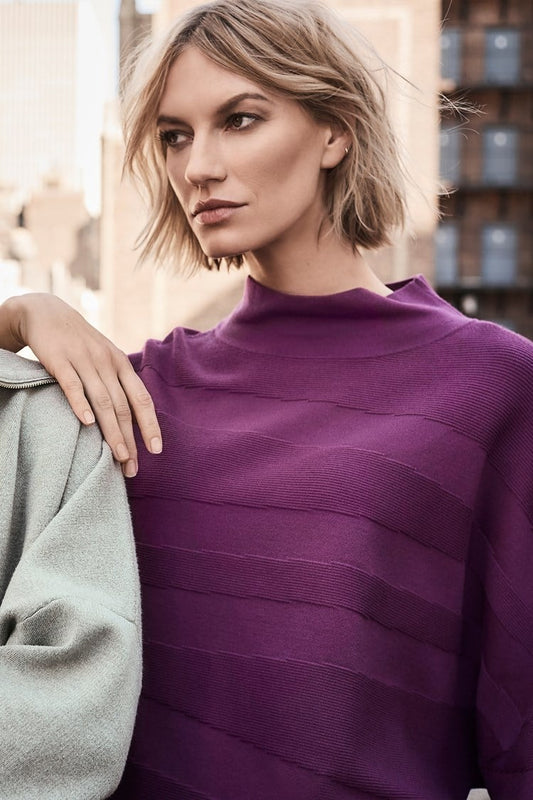 A person with short, tousled blonde hair is wearing a radiant purple *Sweater Knit Mock Neck Boxy Top 243953* by Joseph Ribkoff, which features elegant bell sleeves and horizontal patterns. They rest their arm across the shoulder of another person who is partially visible on the left side of the image under a blurred background of city buildings.