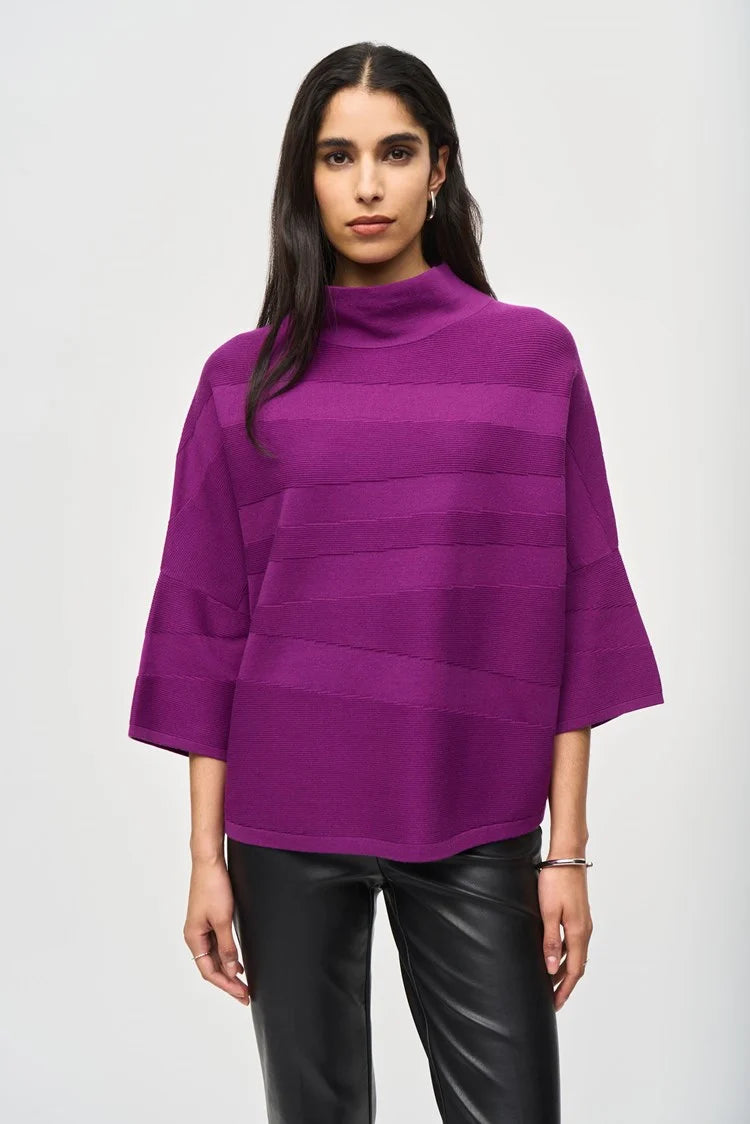 A person with short, tousled blonde hair is wearing a radiant purple *Sweater Knit Mock Neck Boxy Top 243953* by Joseph Ribkoff, which features elegant bell sleeves and horizontal patterns. They rest their arm across the shoulder of another person who is partially visible on the left side of the image under a blurred background of city buildings.