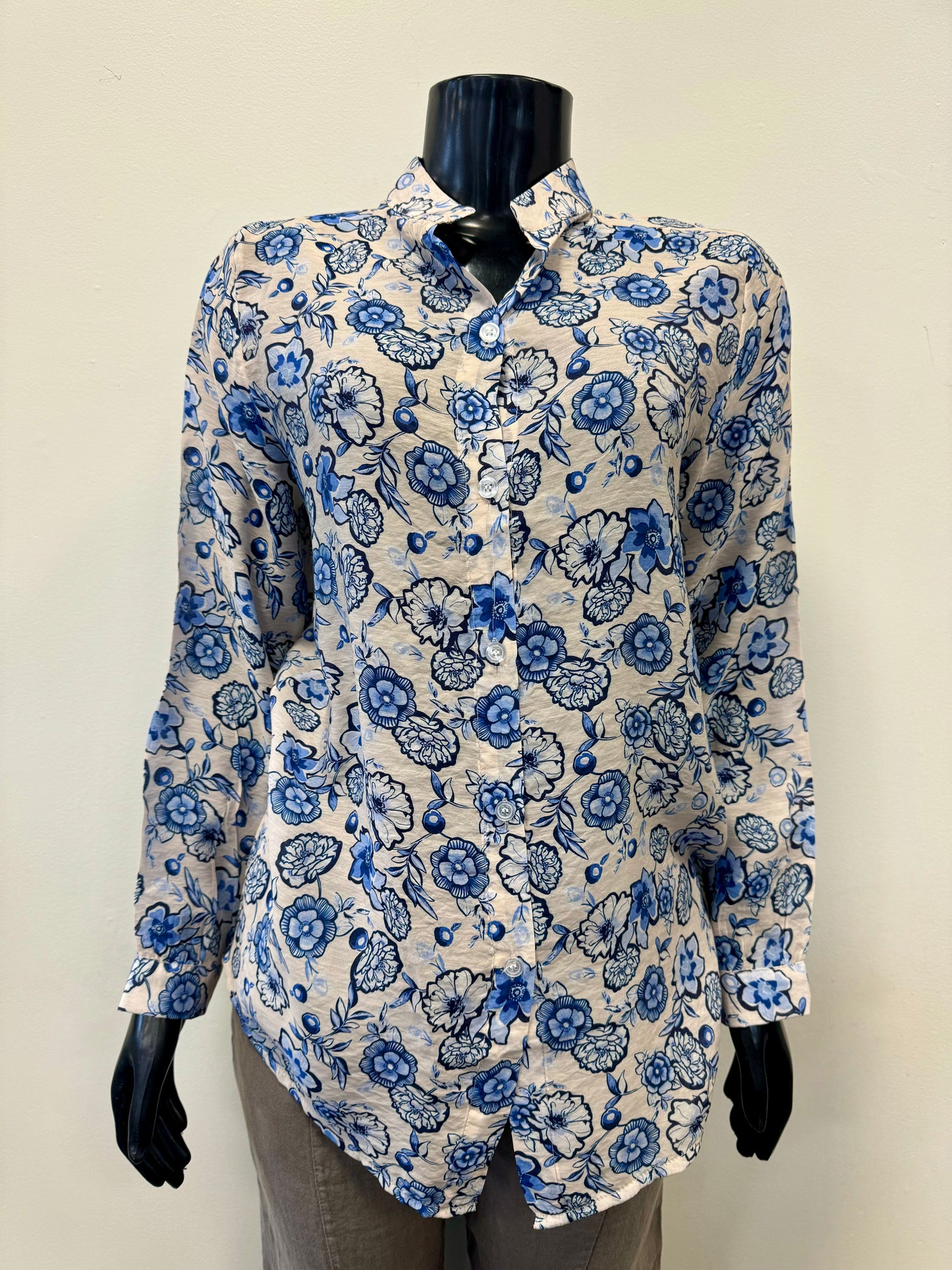 A mannequin dressed in a long-sleeve "Allover Print Blouse - FEMME FATALE T-24B" by Femme Fatale, featuring a blue and white floral pattern, exudes a luxurious feel. The blouse is paired with gray pants, creating an effortlessly chic look. The mannequin is set against a plain, light-colored background.