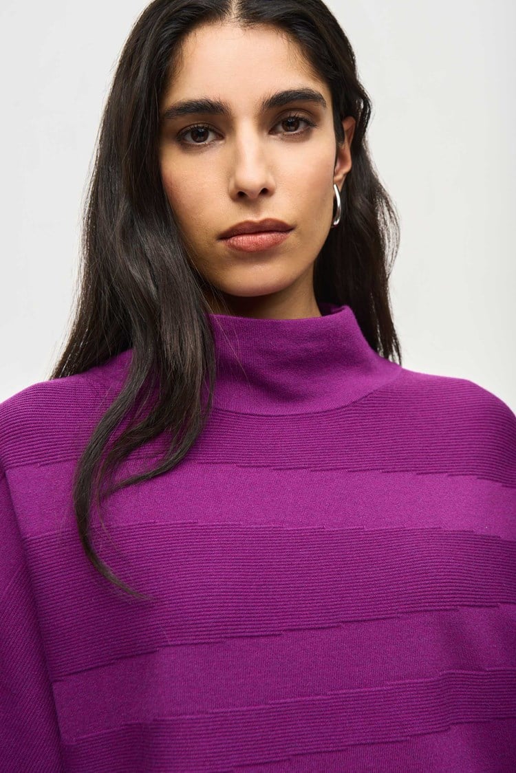 A person with short, tousled blonde hair is wearing a radiant purple *Sweater Knit Mock Neck Boxy Top 243953* by Joseph Ribkoff, which features elegant bell sleeves and horizontal patterns. They rest their arm across the shoulder of another person who is partially visible on the left side of the image under a blurred background of city buildings.