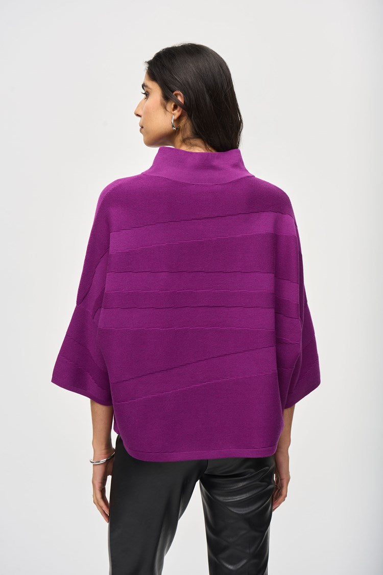 A person with short, tousled blonde hair is wearing a radiant purple *Sweater Knit Mock Neck Boxy Top 243953* by Joseph Ribkoff, which features elegant bell sleeves and horizontal patterns. They rest their arm across the shoulder of another person who is partially visible on the left side of the image under a blurred background of city buildings.