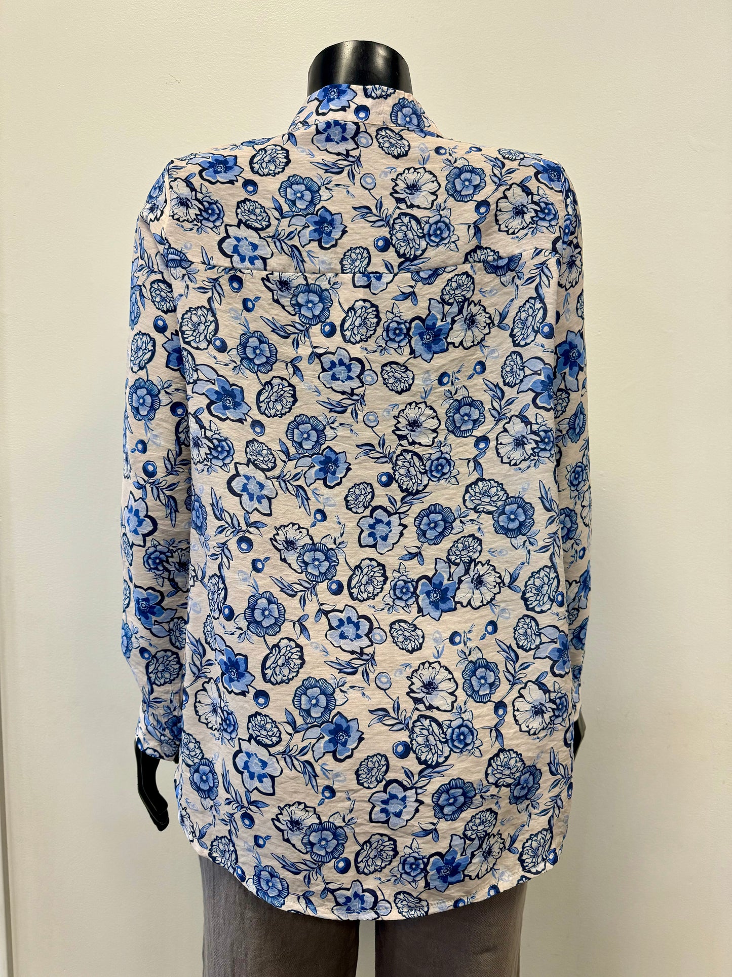 A mannequin dressed in a long-sleeve "Allover Print Blouse - FEMME FATALE T-24B" by Femme Fatale, featuring a blue and white floral pattern, exudes a luxurious feel. The blouse is paired with gray pants, creating an effortlessly chic look. The mannequin is set against a plain, light-colored background.