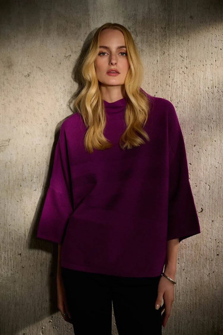 A person with short, tousled blonde hair is wearing a radiant purple *Sweater Knit Mock Neck Boxy Top 243953* by Joseph Ribkoff, which features elegant bell sleeves and horizontal patterns. They rest their arm across the shoulder of another person who is partially visible on the left side of the image under a blurred background of city buildings.