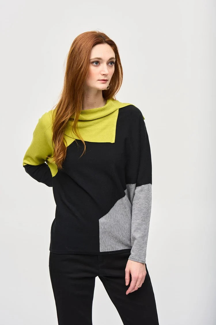 A woman with long red hair stands against a plain background, wearing the Color-Block Jacquard Sweater Top 243941 by Joseph Ribkoff. The cozy fashion sweater in black, gray, and lime green features a unique geometric design with an asymmetrical neckline. She has one hand resting on her hip and is looking to the side.