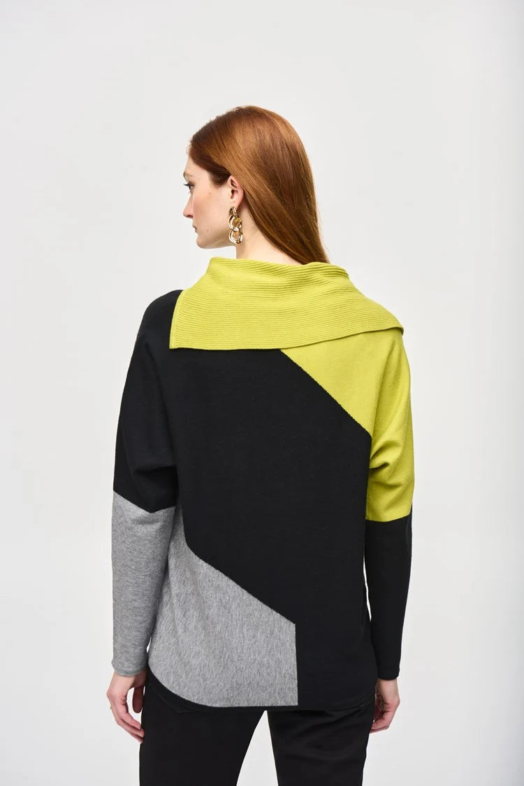 A woman with long red hair stands against a plain background, wearing the Color-Block Jacquard Sweater Top 243941 by Joseph Ribkoff. The cozy fashion sweater in black, gray, and lime green features a unique geometric design with an asymmetrical neckline. She has one hand resting on her hip and is looking to the side.