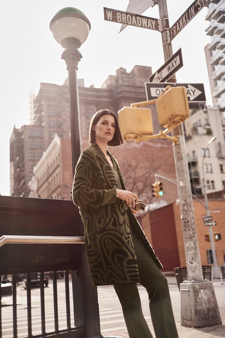 A woman with straight brown hair stands on a city street corner, next to a street sign for W Broadway and Franklin St. She is wearing the Embossed Jacquard Knit Cover Up 243932 by Joseph Ribkoff along with matching pants. The background features tall buildings, traffic lights, and a pedestrian subway entrance.