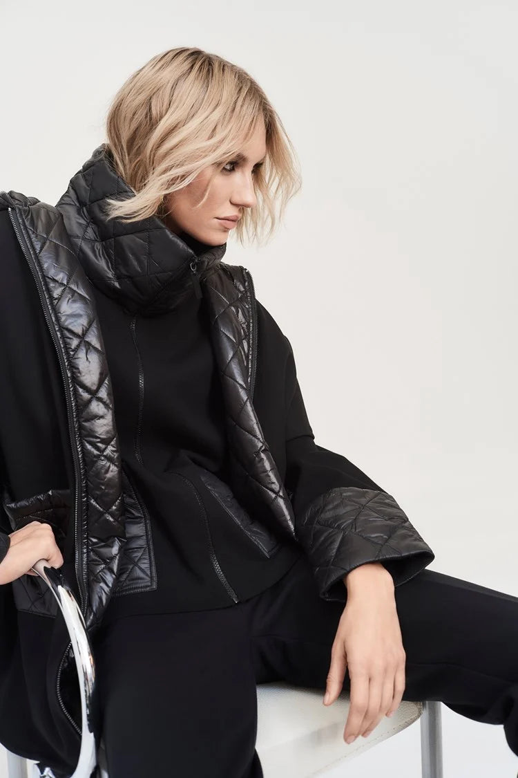 A woman with short blonde hair poses against a plain background, donning the Sweater Knit Zipped Jacket 243933 by Joseph Ribkoff. The black jacket features quilted nylon detailing on the collar, sleeves, and sides. With her hands placed inside the open sides of its front zipper closure, she gazes confidently at the camera.