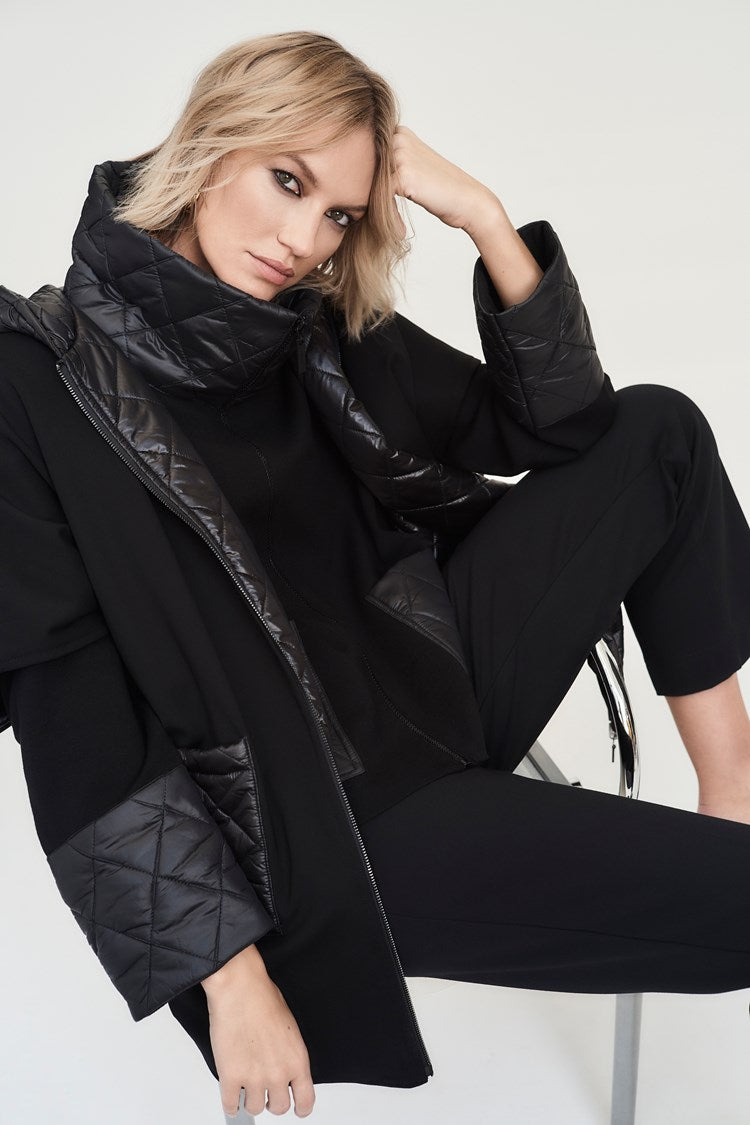 A person with short blonde hair sits on a chair, slightly leaning forward with an elbow resting on their knee and their hand supporting their head. They are adorned in the stylish Heavy Knit Reversible Hooded Cape by Joseph Ribkoff, which is made from quality heavy knit fabric and features a large quilted collar and matching cuffs, paired with black pants.