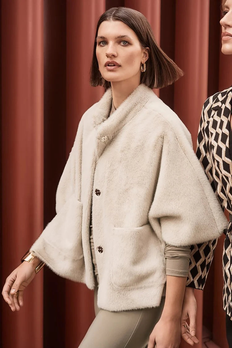 A woman with a bob haircut wears the light-colored, oversized **Bonded Faux Fur Suede Jacket 243917 by Joseph Ribkoff** over a mock neck top and beige pants, walking next to another person. The background features vertical red-brown stripes.