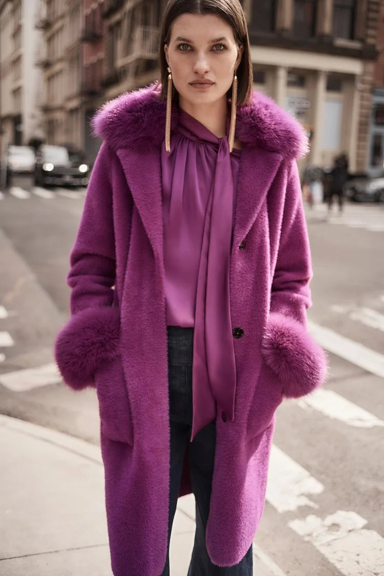 A woman stands on a city street wearing the Joseph Ribkoff Feather Yarn and Faux Fur Sweater Coat 243923 in a vibrant purple color, layered over a matching blouse. She has shoulder-length brown hair, is looking directly at the camera, and the background shows buildings and cars at an intersection.