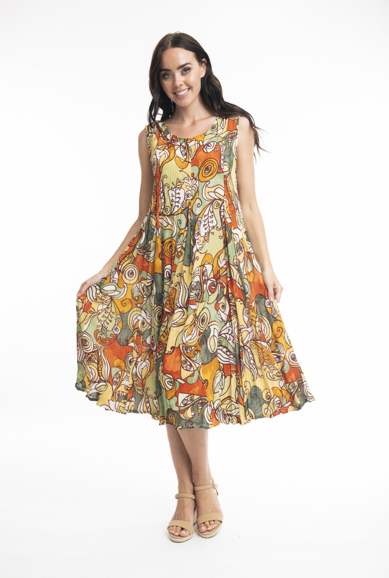 A woman with long dark hair is smiling as she poses against a white background, holding out the sides of her ORIENTIQUE Ephesus Dress Godet Sleeveless Style: 3067. The sustainable rayon, knee-length dress showcases a vibrant, multicolored abstract pattern. She completes the look with beige wedges.