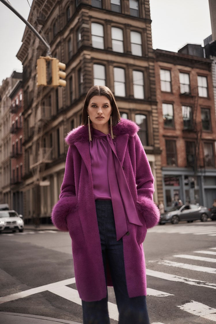 A woman stands on a city street wearing the Joseph Ribkoff Feather Yarn and Faux Fur Sweater Coat 243923 in a vibrant purple color, layered over a matching blouse. She has shoulder-length brown hair, is looking directly at the camera, and the background shows buildings and cars at an intersection.