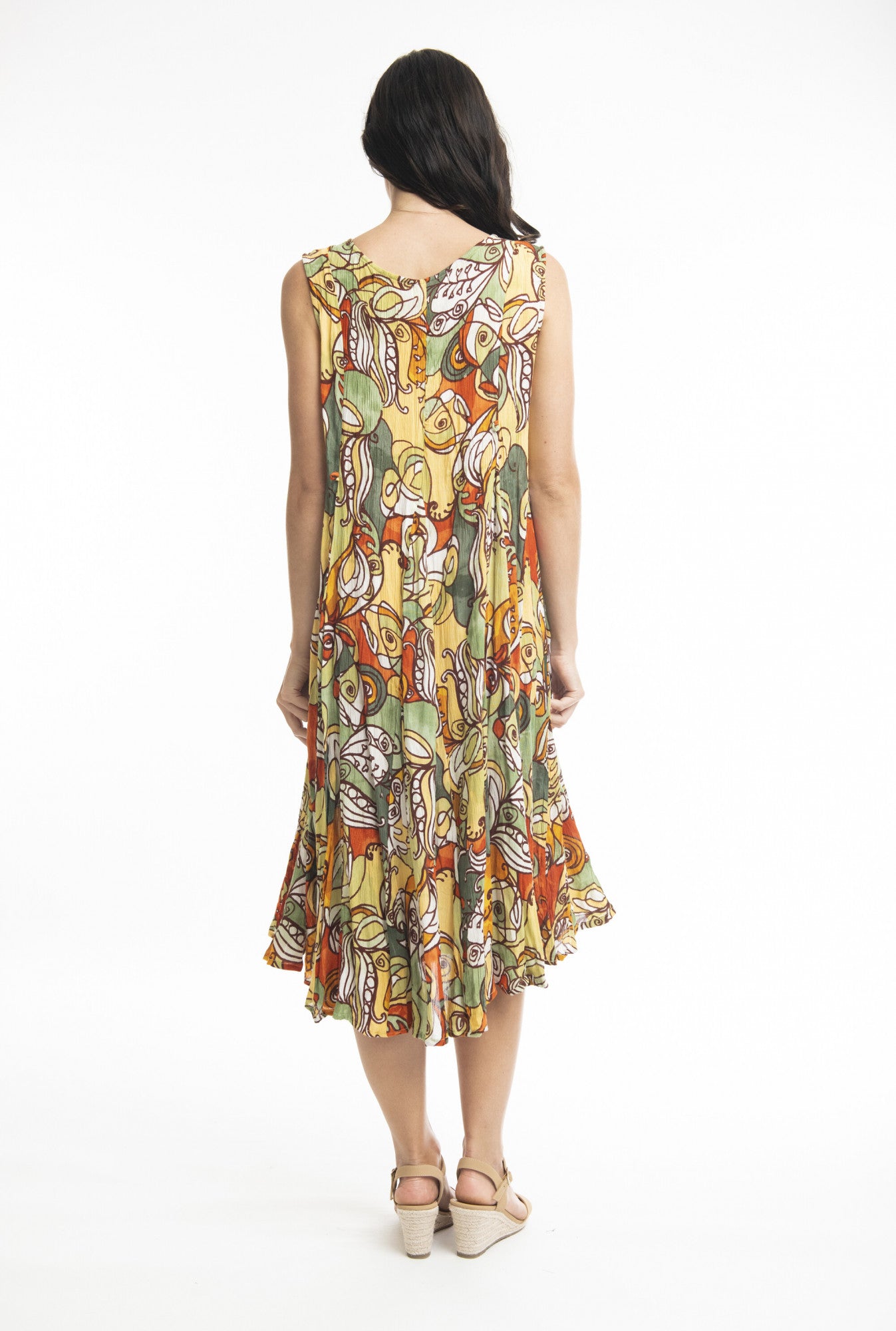 A woman with long dark hair is smiling as she poses against a white background, holding out the sides of her ORIENTIQUE Ephesus Dress Godet Sleeveless Style: 3067. The sustainable rayon, knee-length dress showcases a vibrant, multicolored abstract pattern. She completes the look with beige wedges.