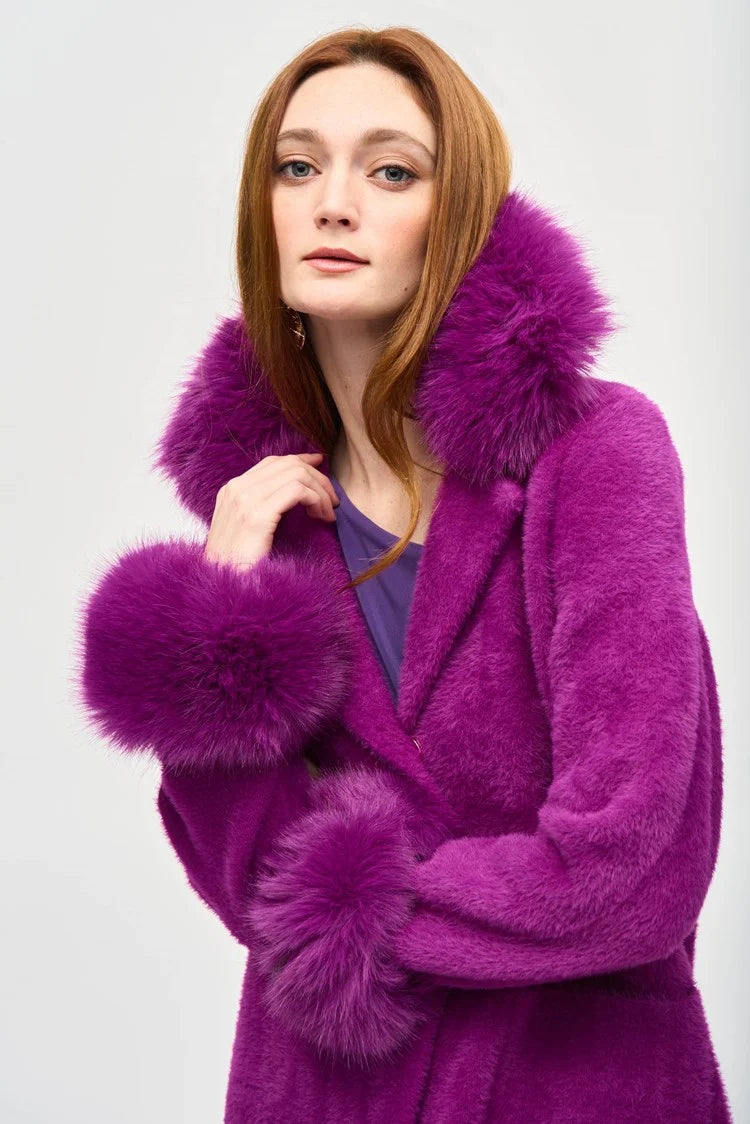 A woman stands on a city street wearing the Joseph Ribkoff Feather Yarn and Faux Fur Sweater Coat 243923 in a vibrant purple color, layered over a matching blouse. She has shoulder-length brown hair, is looking directly at the camera, and the background shows buildings and cars at an intersection.