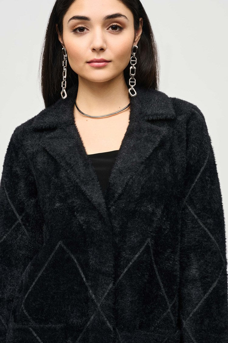 A person stands against a light gray background, wearing the Joseph Ribkoff Notched Collar Coat (style 233951F24) in black over a black top and black pants. They also have on black heeled ankle boots and large, dangling earrings. Their hands are relaxed at their sides.