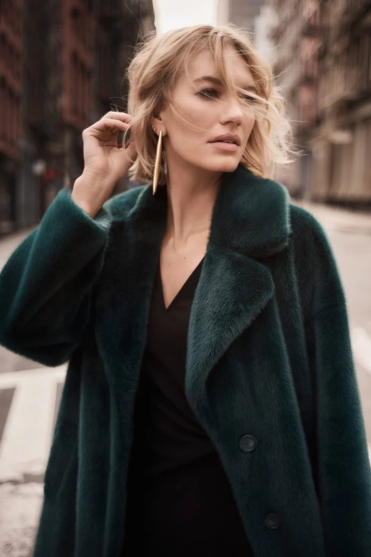 A woman with short blonde hair stands on a city street, exuding chic style in the Joseph Ribkoff Faux Fur Straight Coat 243902. The dark emerald green coat, featuring a notched collar, is draped over a black top. She gazes off to the side, the wind tousling her hair and revealing large gold hoop earrings.