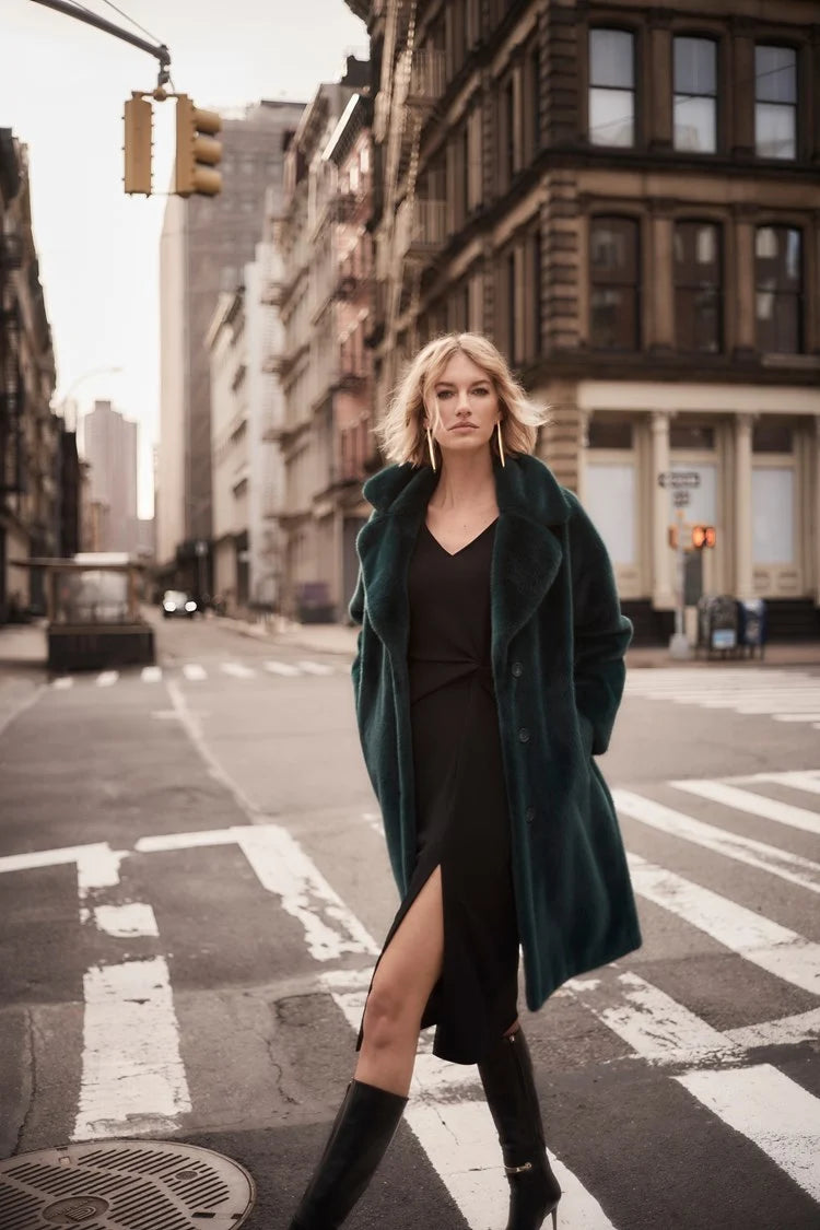 A woman with short blonde hair stands on a city street, exuding chic style in the Joseph Ribkoff Faux Fur Straight Coat 243902. The dark emerald green coat, featuring a notched collar, is draped over a black top. She gazes off to the side, the wind tousling her hair and revealing large gold hoop earrings.