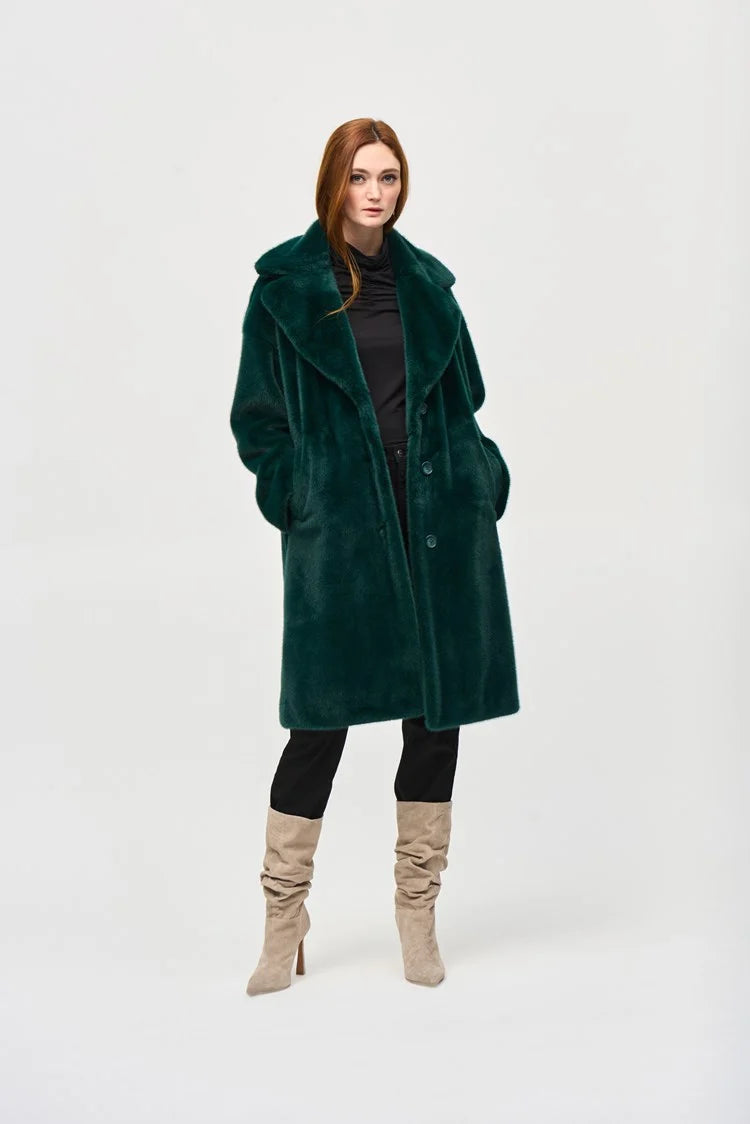 A woman with short blonde hair stands on a city street, exuding chic style in the Joseph Ribkoff Faux Fur Straight Coat 243902. The dark emerald green coat, featuring a notched collar, is draped over a black top. She gazes off to the side, the wind tousling her hair and revealing large gold hoop earrings.