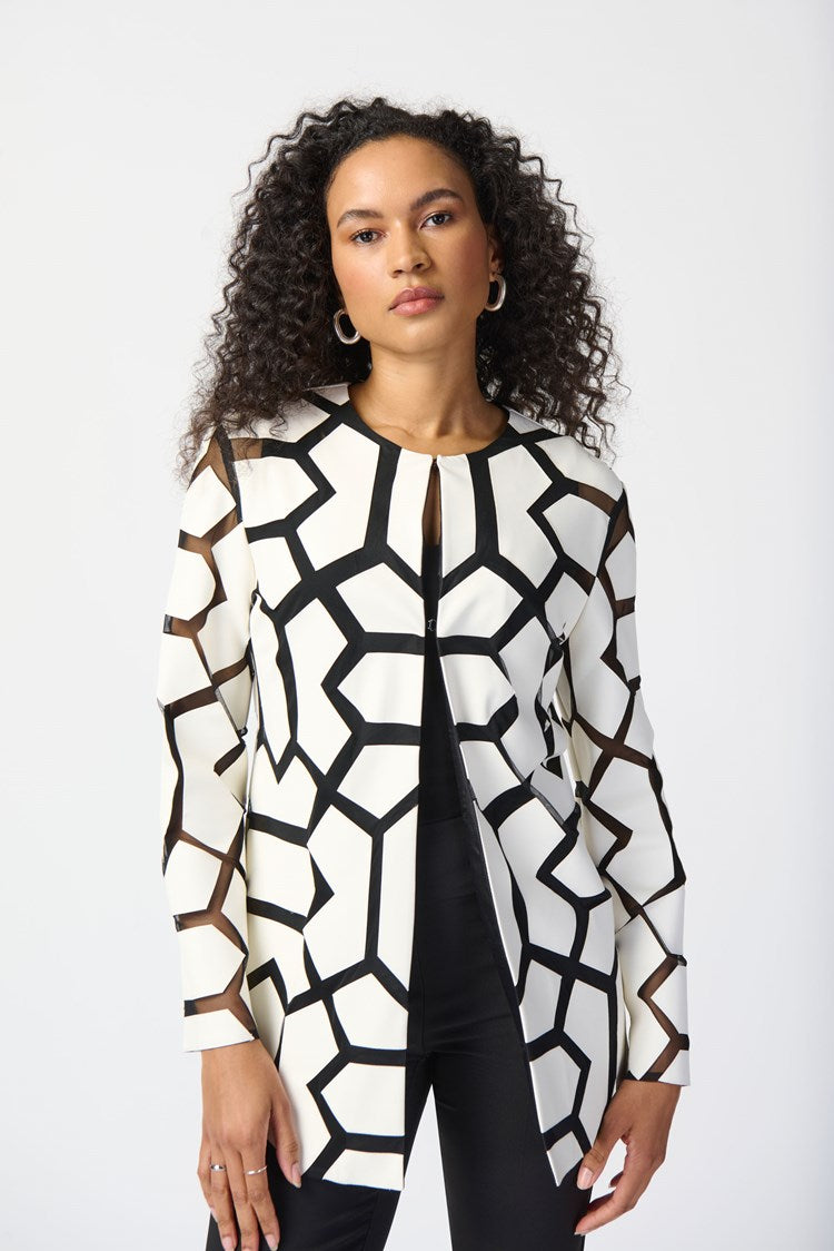 A woman with curly hair stands against a plain white background. She is wearing a white top with a bold black geometric pattern, black pants, and sleek black high-heeled sandals. Draped over her shoulders is the Joseph Ribkoff Laser-Cut Leatherette on Mesh Jacket 241905, enhancing her confident and professional demeanor with a contemporary aesthetic.