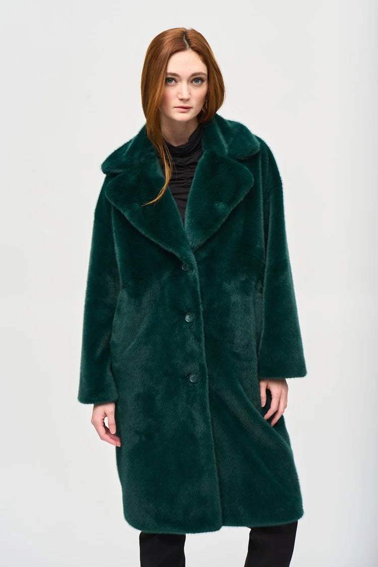 A woman with short blonde hair stands on a city street, exuding chic style in the Joseph Ribkoff Faux Fur Straight Coat 243902. The dark emerald green coat, featuring a notched collar, is draped over a black top. She gazes off to the side, the wind tousling her hair and revealing large gold hoop earrings.