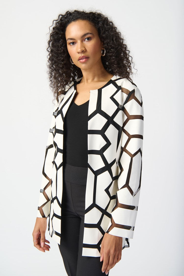 A woman with curly hair stands against a plain white background. She is wearing a white top with a bold black geometric pattern, black pants, and sleek black high-heeled sandals. Draped over her shoulders is the Joseph Ribkoff Laser-Cut Leatherette on Mesh Jacket 241905, enhancing her confident and professional demeanor with a contemporary aesthetic.