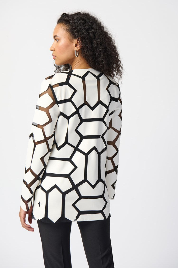 A woman with curly hair stands against a plain white background. She is wearing a white top with a bold black geometric pattern, black pants, and sleek black high-heeled sandals. Draped over her shoulders is the Joseph Ribkoff Laser-Cut Leatherette on Mesh Jacket 241905, enhancing her confident and professional demeanor with a contemporary aesthetic.