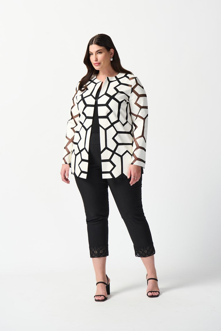 A woman with curly hair stands against a plain white background. She is wearing a white top with a bold black geometric pattern, black pants, and sleek black high-heeled sandals. Draped over her shoulders is the Joseph Ribkoff Laser-Cut Leatherette on Mesh Jacket 241905, enhancing her confident and professional demeanor with a contemporary aesthetic.