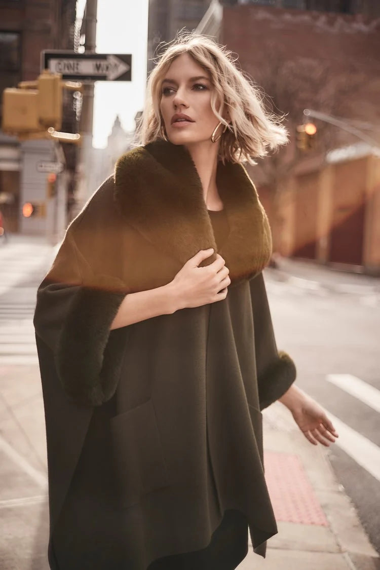 A stylish woman with short, wavy blonde hair is walking on a city street. She is wearing a fashionable Joseph Ribkoff dark green coat with faux fur trim, layered over the chic Brushed Jacquard and Faux Fur Cape 243930 by Joseph Ribkoff. The background features an urban setting with buildings, traffic lights, and a one-way street sign. The sun casts a warm glow.