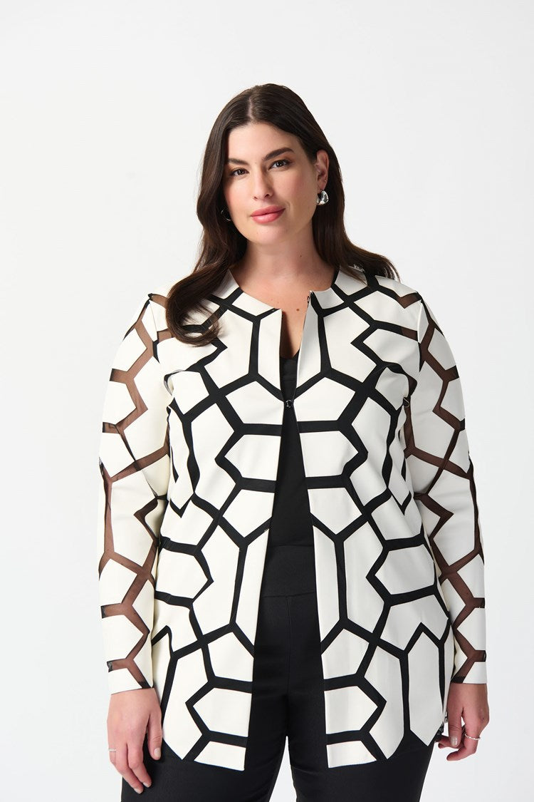 A woman with curly hair stands against a plain white background. She is wearing a white top with a bold black geometric pattern, black pants, and sleek black high-heeled sandals. Draped over her shoulders is the Joseph Ribkoff Laser-Cut Leatherette on Mesh Jacket 241905, enhancing her confident and professional demeanor with a contemporary aesthetic.