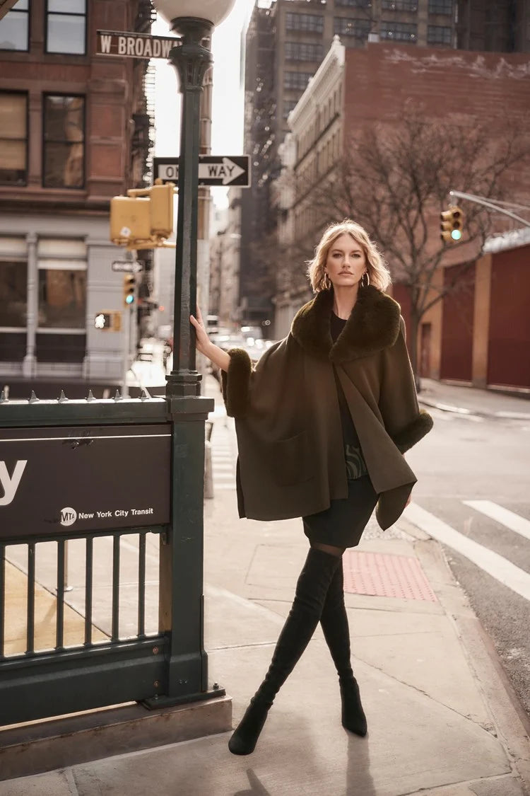 A stylish woman with short, wavy blonde hair is walking on a city street. She is wearing a fashionable Joseph Ribkoff dark green coat with faux fur trim, layered over the chic Brushed Jacquard and Faux Fur Cape 243930 by Joseph Ribkoff. The background features an urban setting with buildings, traffic lights, and a one-way street sign. The sun casts a warm glow.