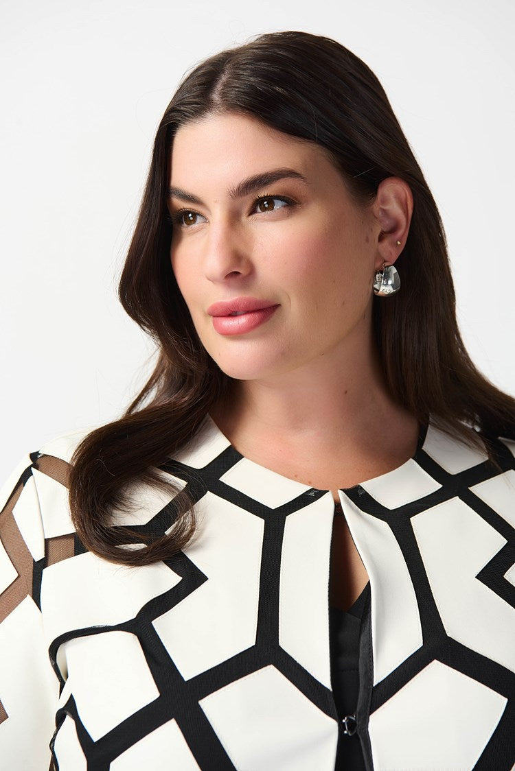 A woman with curly hair stands against a plain white background. She is wearing a white top with a bold black geometric pattern, black pants, and sleek black high-heeled sandals. Draped over her shoulders is the Joseph Ribkoff Laser-Cut Leatherette on Mesh Jacket 241905, enhancing her confident and professional demeanor with a contemporary aesthetic.