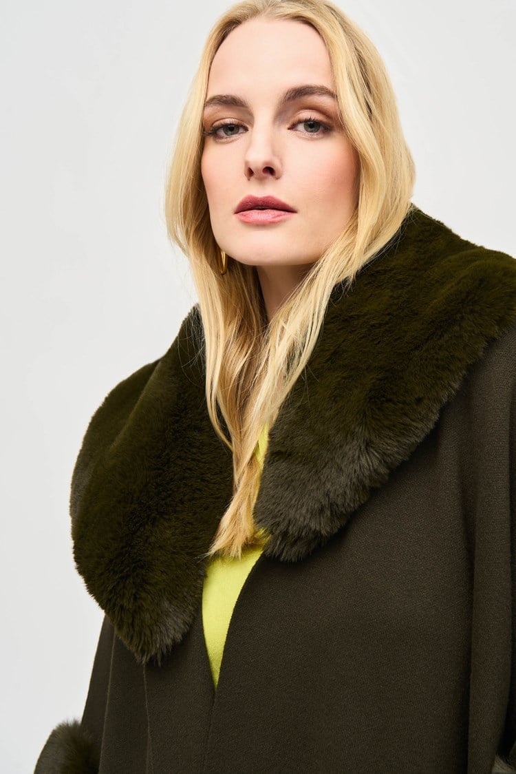 A stylish woman with short, wavy blonde hair is walking on a city street. She is wearing a fashionable Joseph Ribkoff dark green coat with faux fur trim, layered over the chic Brushed Jacquard and Faux Fur Cape 243930 by Joseph Ribkoff. The background features an urban setting with buildings, traffic lights, and a one-way street sign. The sun casts a warm glow.