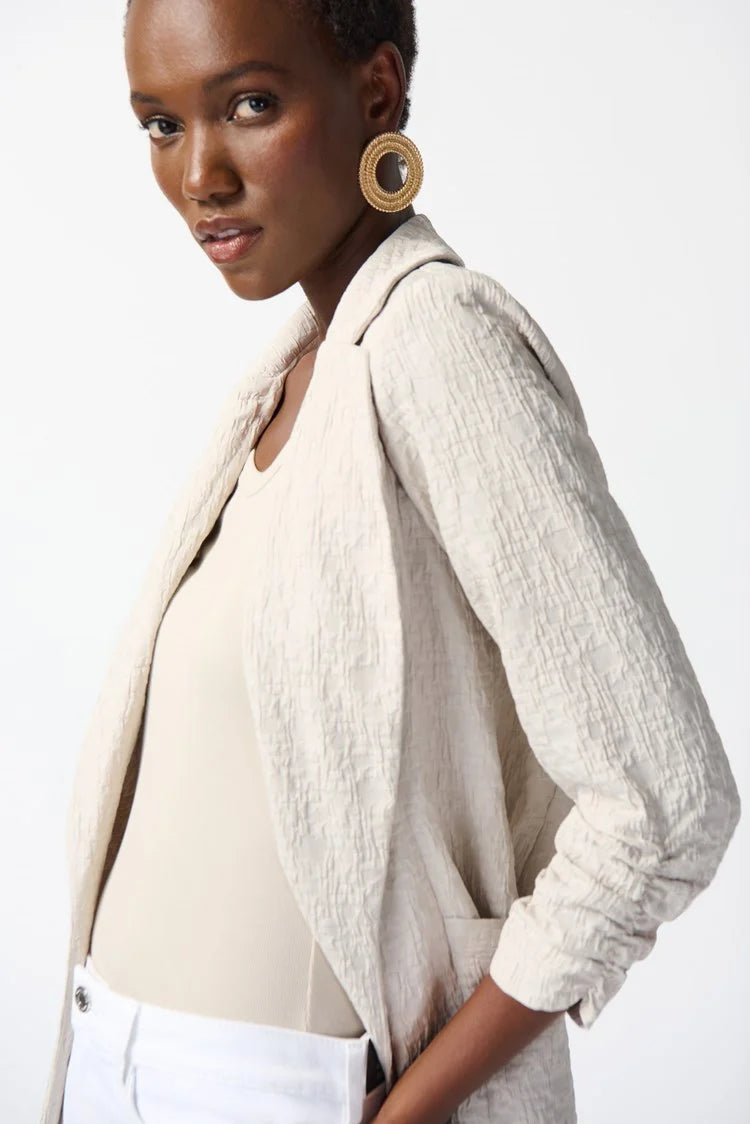 A person with short hair wears a light beige Woven Jacquard Boxy Blazer 242034 from Joseph Ribkoff, featuring a textured finish, over a matching top and white pants. One hand is in the pants pocket. They also wear large, circular gold earrings and have a confident expression, posing against a plain white background.