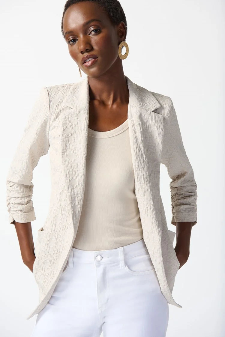 A person with short hair wears a light beige Woven Jacquard Boxy Blazer 242034 from Joseph Ribkoff, featuring a textured finish, over a matching top and white pants. One hand is in the pants pocket. They also wear large, circular gold earrings and have a confident expression, posing against a plain white background.