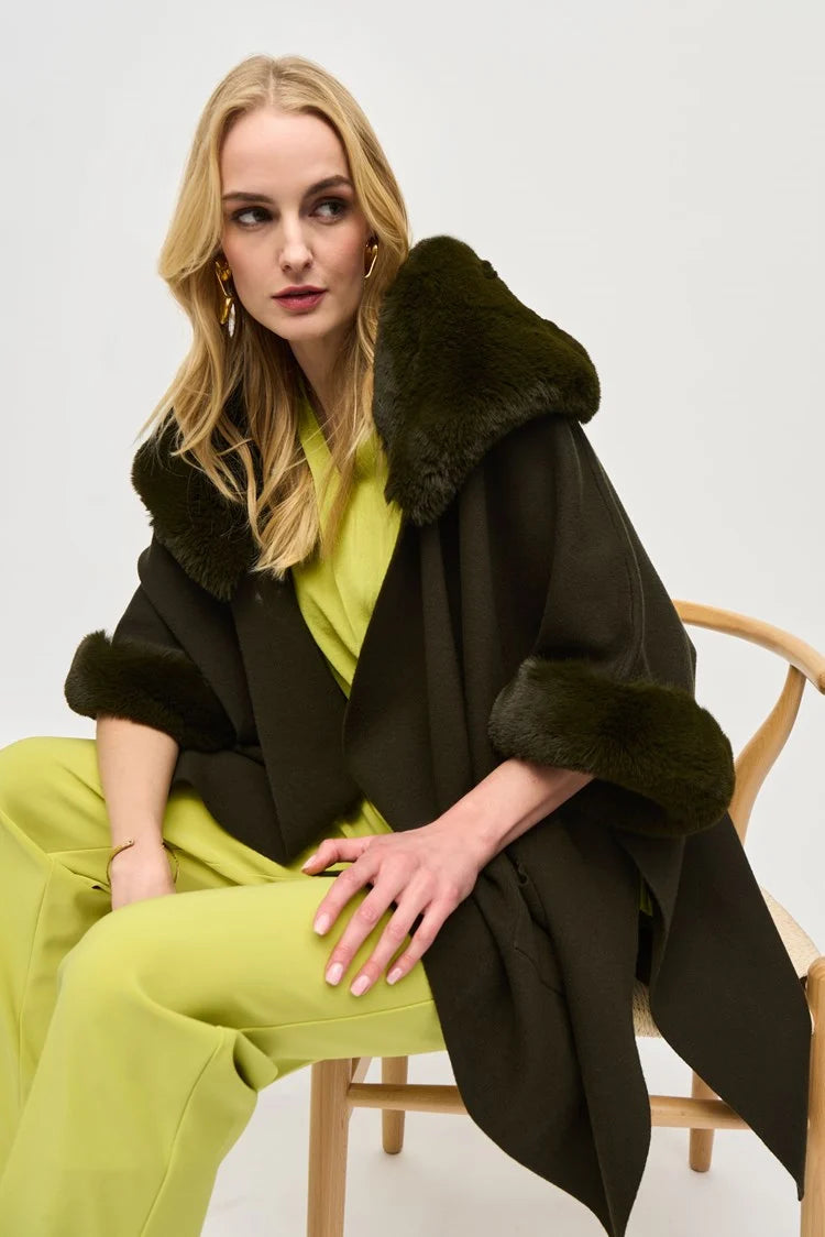 A stylish woman with short, wavy blonde hair is walking on a city street. She is wearing a fashionable Joseph Ribkoff dark green coat with faux fur trim, layered over the chic Brushed Jacquard and Faux Fur Cape 243930 by Joseph Ribkoff. The background features an urban setting with buildings, traffic lights, and a one-way street sign. The sun casts a warm glow.