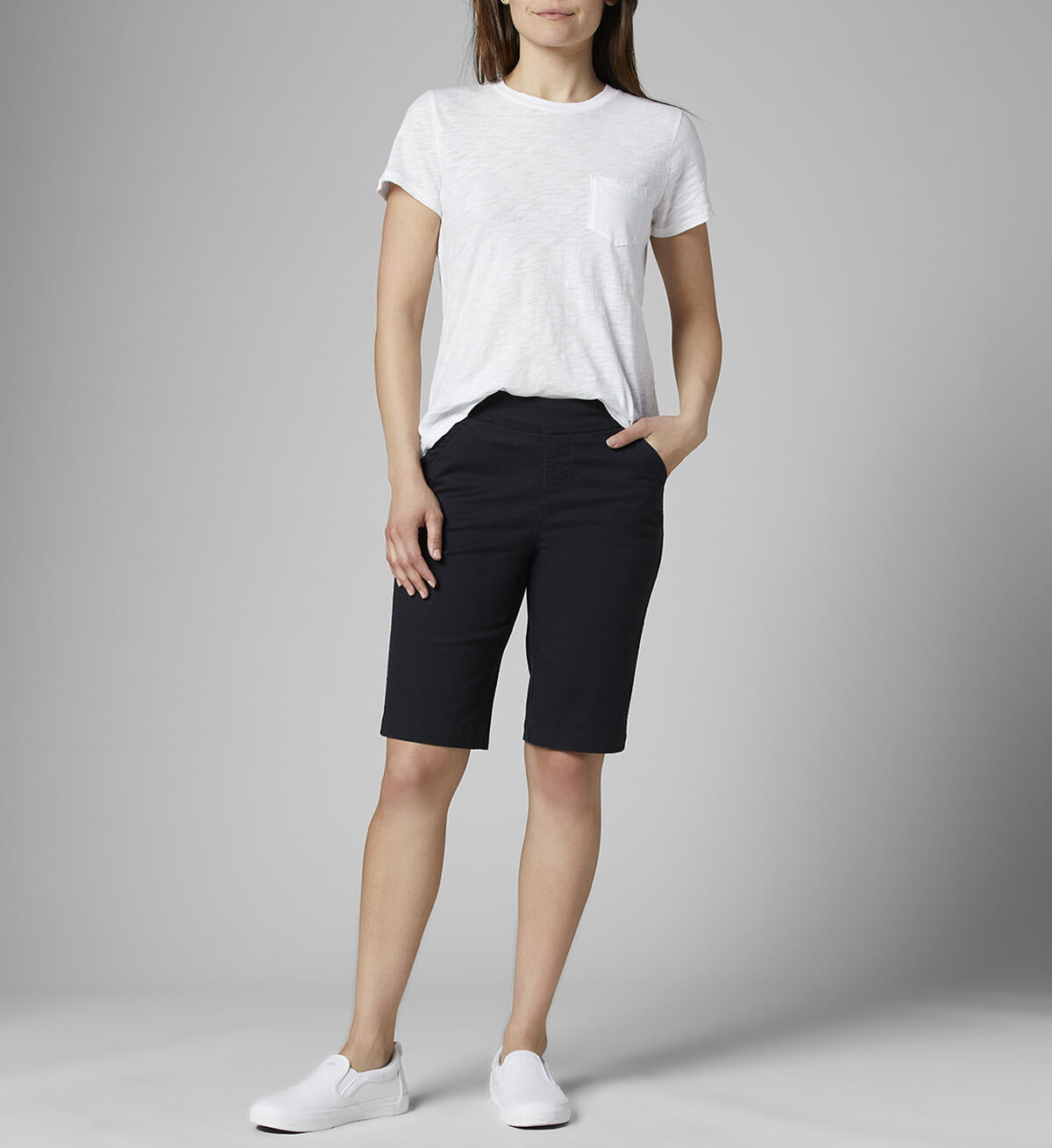 A person stands against a gray background, wearing a white short-sleeved t-shirt with a small pocket on the left side and black JAG Gracie Bermuda-Shorts featuring a pull-on waistband for comfort stretch. They have their right hand in their pocket and wear white slip-on shoes. The person's head is not visible in the image.