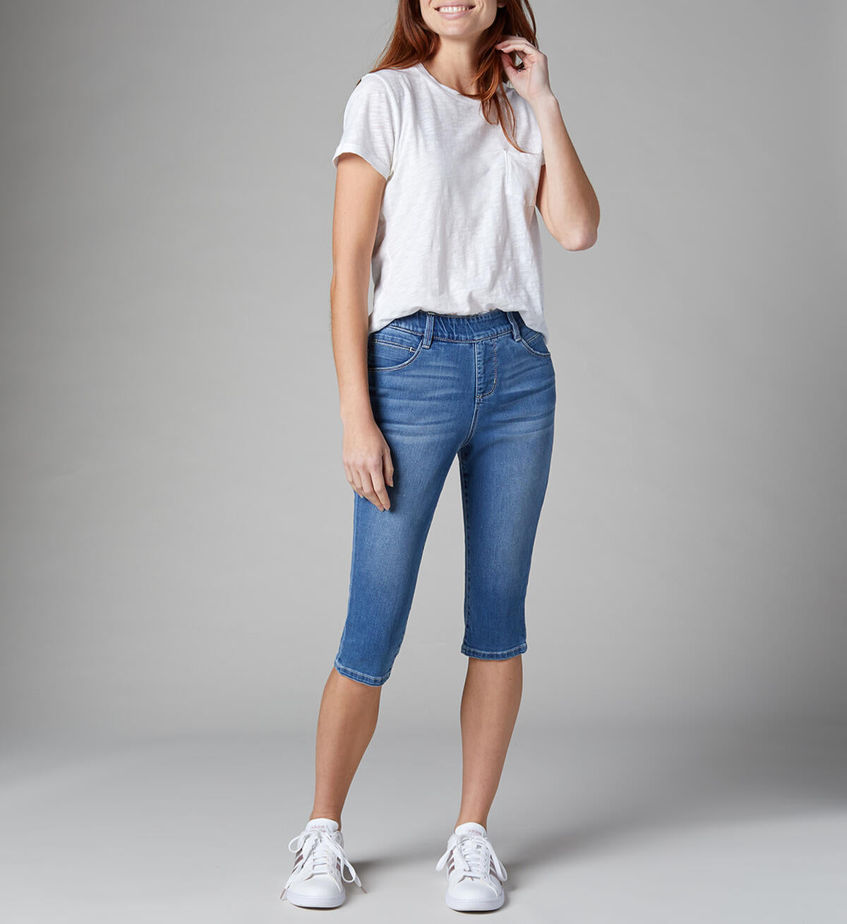 A woman is standing against a plain gray background, wearing a casual white t-shirt, high rise pull-on Bryn Pedal Pusher-Jeans from JAG, and white sneakers. She is smiling, with her right hand touching her hair and her left hand hanging by her side.