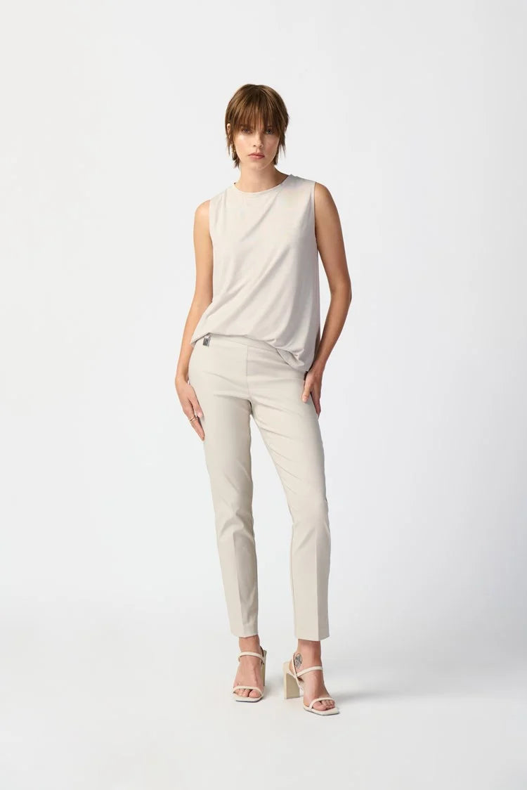 A person with short brown hair is standing against a plain white background. They are wearing a Bamboo Blend Sleeveless Jersey 241289 by Joseph Ribkoff, light gray slim-fit pants, and beige open-toe high-heeled shoes. Their right hand is in their pocket, and their left hand rests by their side.